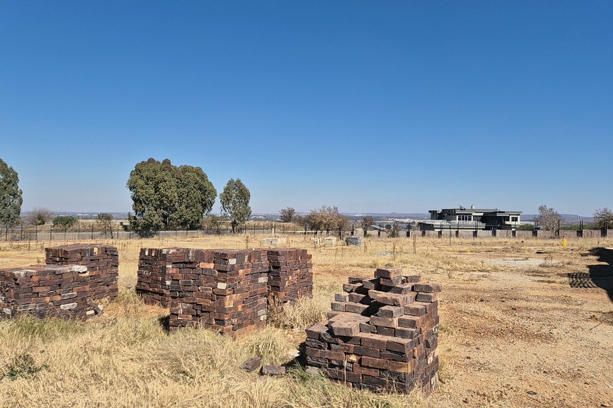 A vacant residential plot featuring dry grass, stacks of construction bricks, and perimeter fencing with distant neighborhood views.