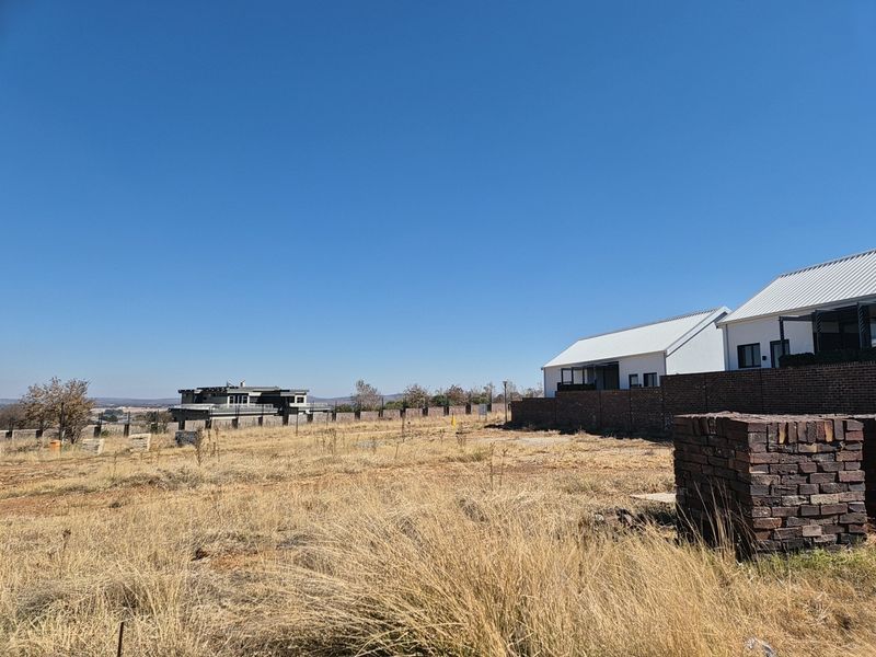 A view of a vacant residential plot featuring dry grass, a brick boundary wall, and a stack of bricks on site.