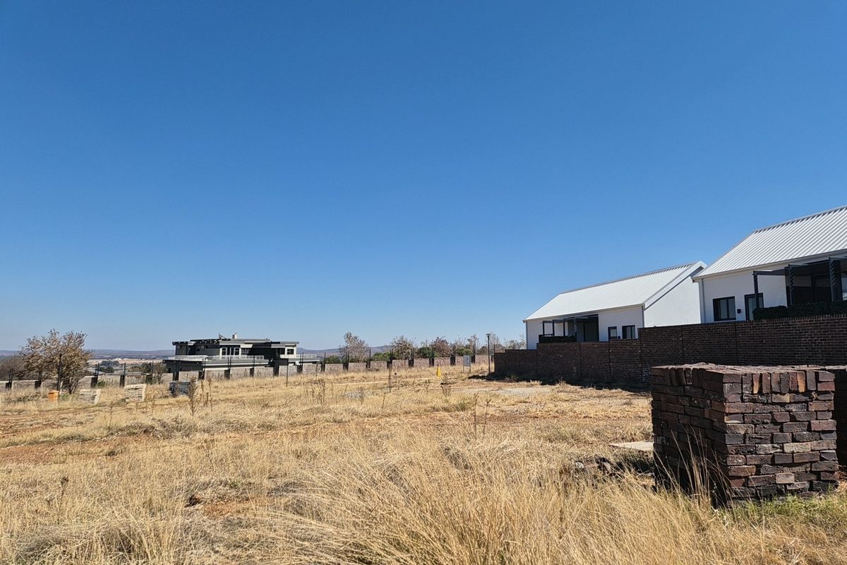 A view of a vacant residential plot featuring dry grass, a brick boundary wall, and a stack of bricks on site.