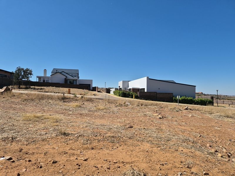A view of a vacant residential plot featuring a paved road and surrounding modern residential architecture in a developed estate setting.
