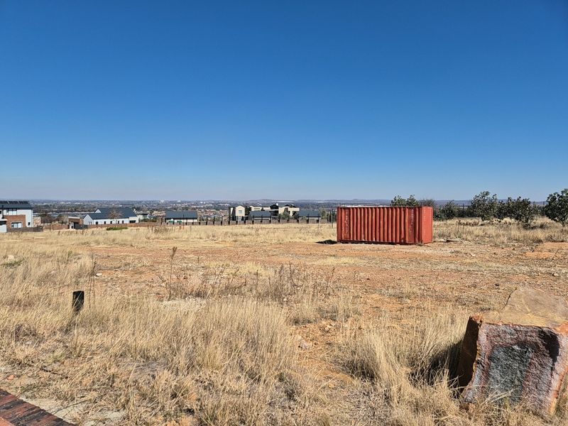 A wide-angle view of a vacant residential plot featuring dry grass, natural terrain, and distant suburban housing developments.