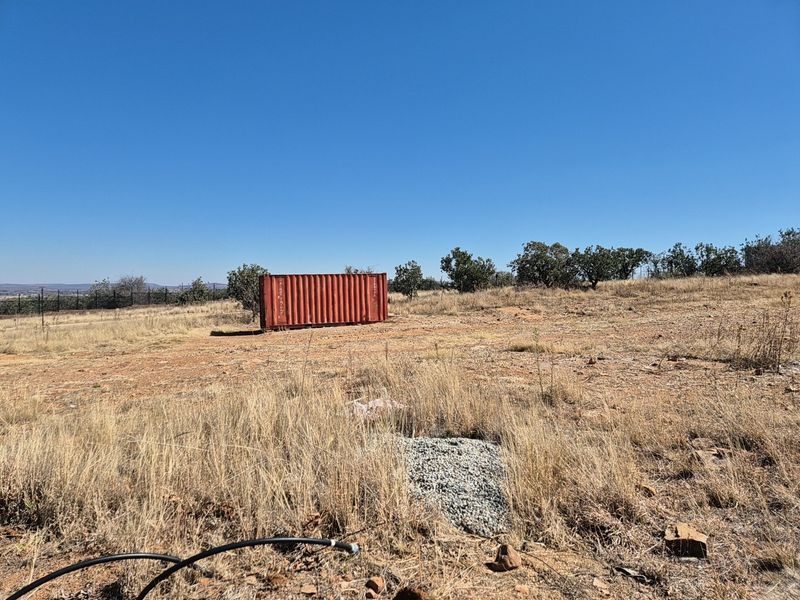 A view of a vacant residential plot featuring dry grass, perimeter fencing, and a shipping container on site.