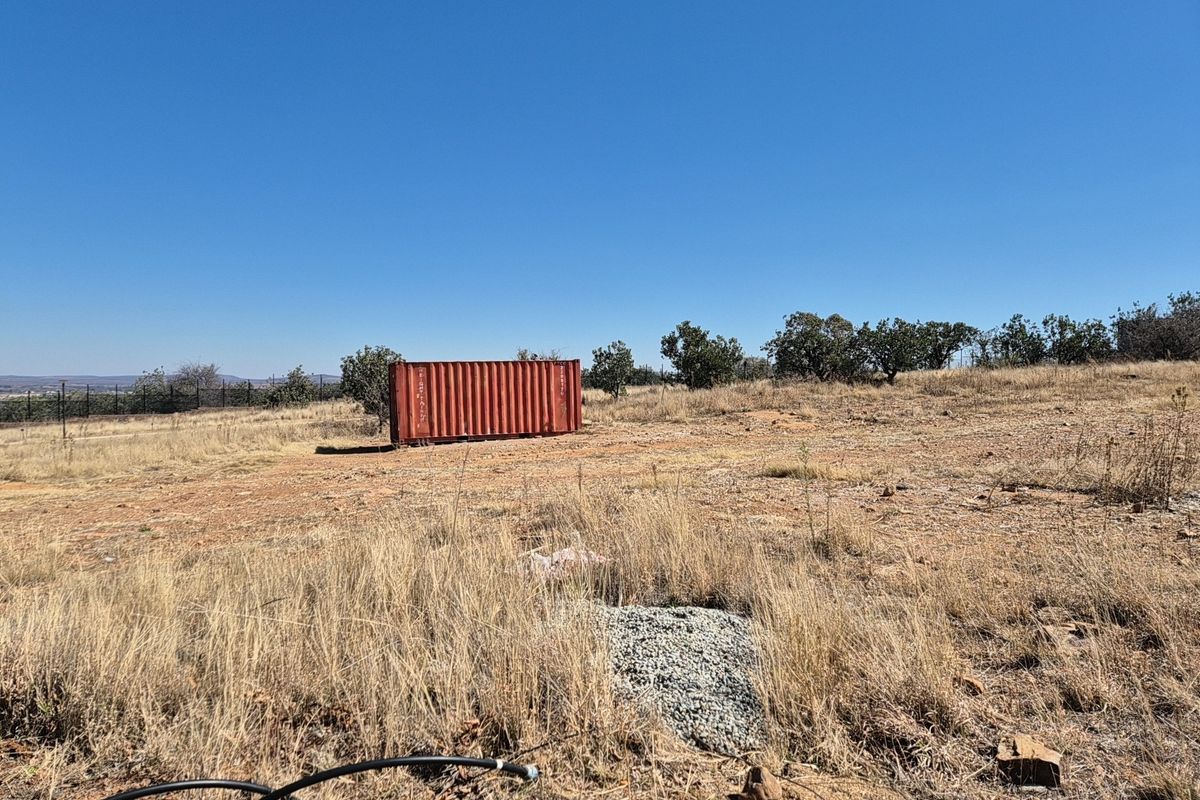 A view of a vacant residential plot featuring dry grass, perimeter fencing, and a shipping container on site.