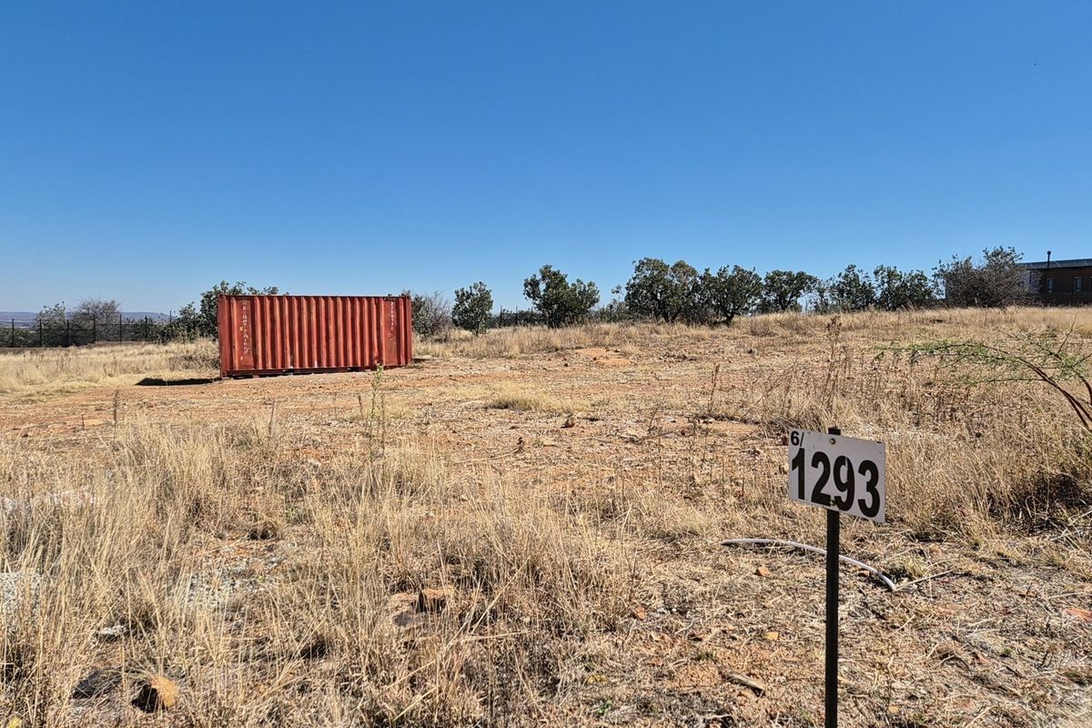 A view of a vacant residential plot featuring dry grass, boundary fencing, and a storage container on site.