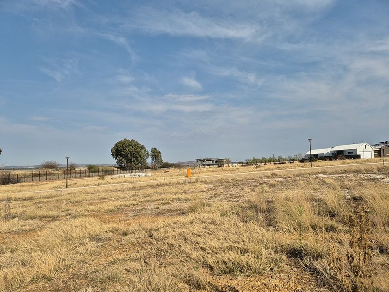 A view of a vacant residential plot featuring dry grass, perimeter fencing, and street lighting in a developing estate.