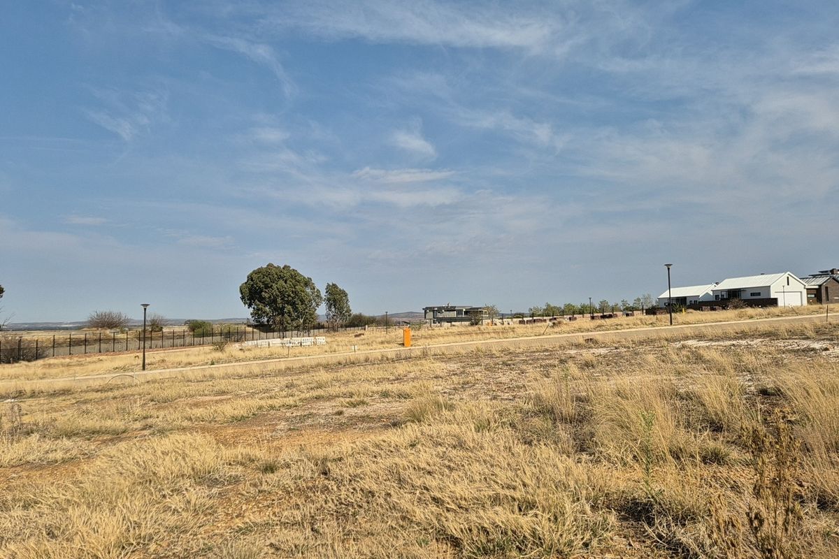A view of a vacant residential plot featuring dry grass, perimeter fencing, and street lighting in a developing estate.