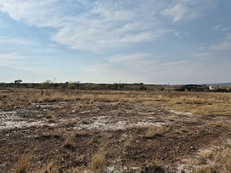 A wide-angle view of a vacant residential plot featuring dry grass, perimeter fencing, and distant neighborhood infrastructure.
