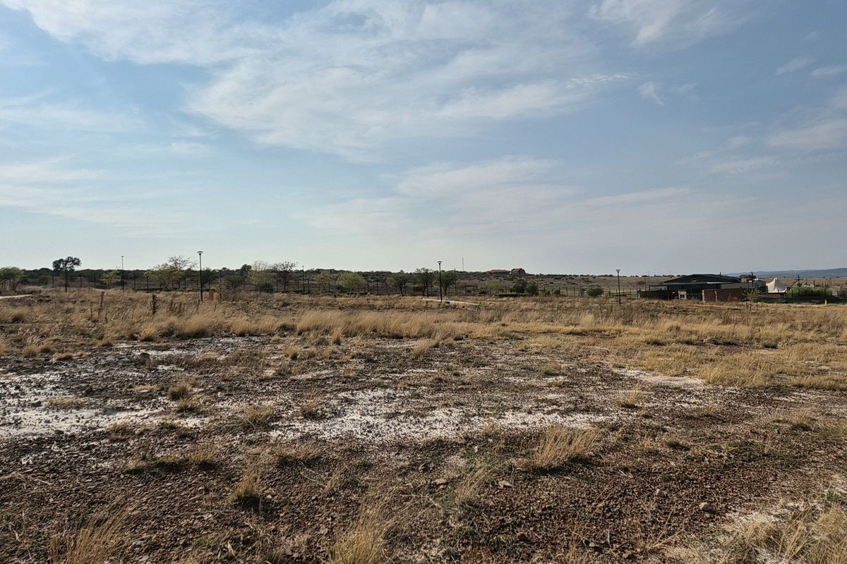A wide-angle view of a vacant residential plot featuring dry grass, perimeter fencing, and distant neighborhood infrastructure.
