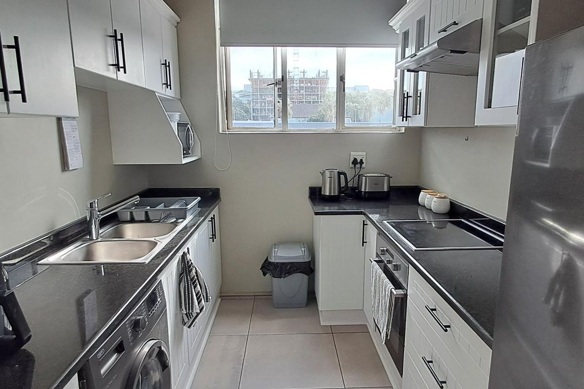 A functional galley-style kitchen featuring white cabinetry, dark stone countertops, a built-in oven, hob, extractor fan, and integrated plumbing for laundry appliances.