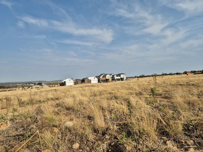 A wide-angle view of a vacant residential plot featuring dry grass terrain with established modern homes visible in the background.