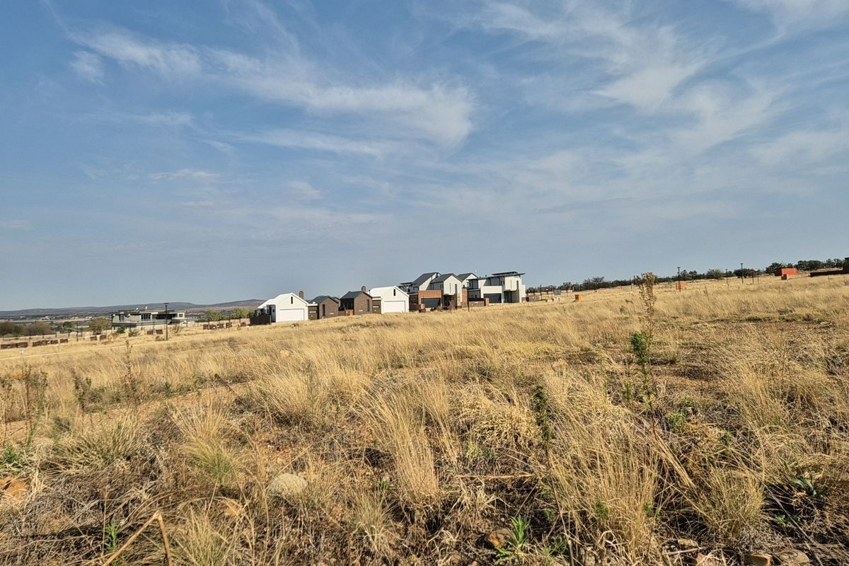 A wide-angle view of a vacant residential plot featuring dry grass terrain with established modern homes visible in the background.