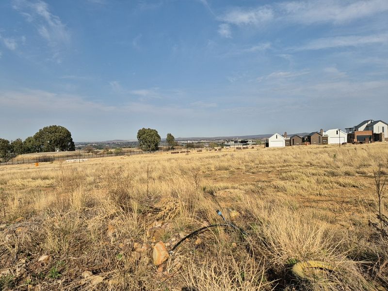 A wide-angle view of a vacant residential plot featuring dry grass, perimeter fencing, and distant neighborhood houses under a clear blue sky.