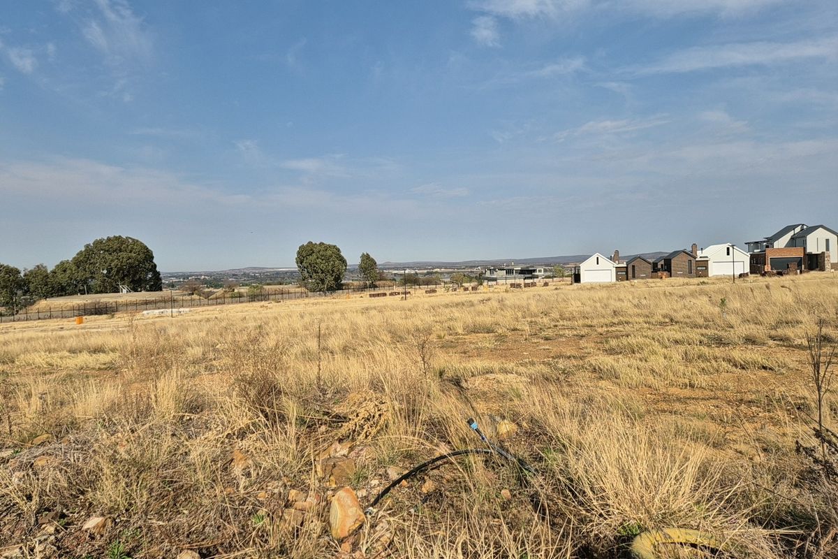A wide-angle view of a vacant residential plot featuring dry grass, perimeter fencing, and distant neighborhood houses under a clear blue sky.