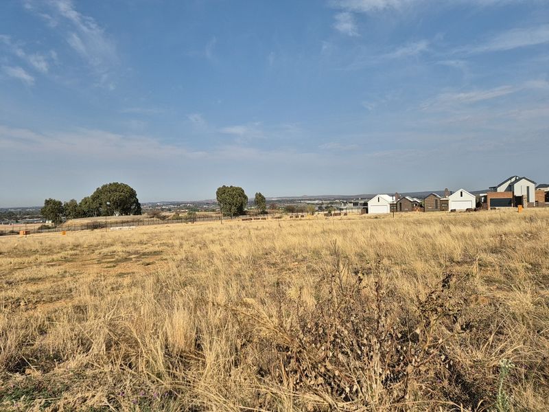 A wide-angle view of a vacant residential plot featuring dry grass and surrounding neighborhood development in the distance.