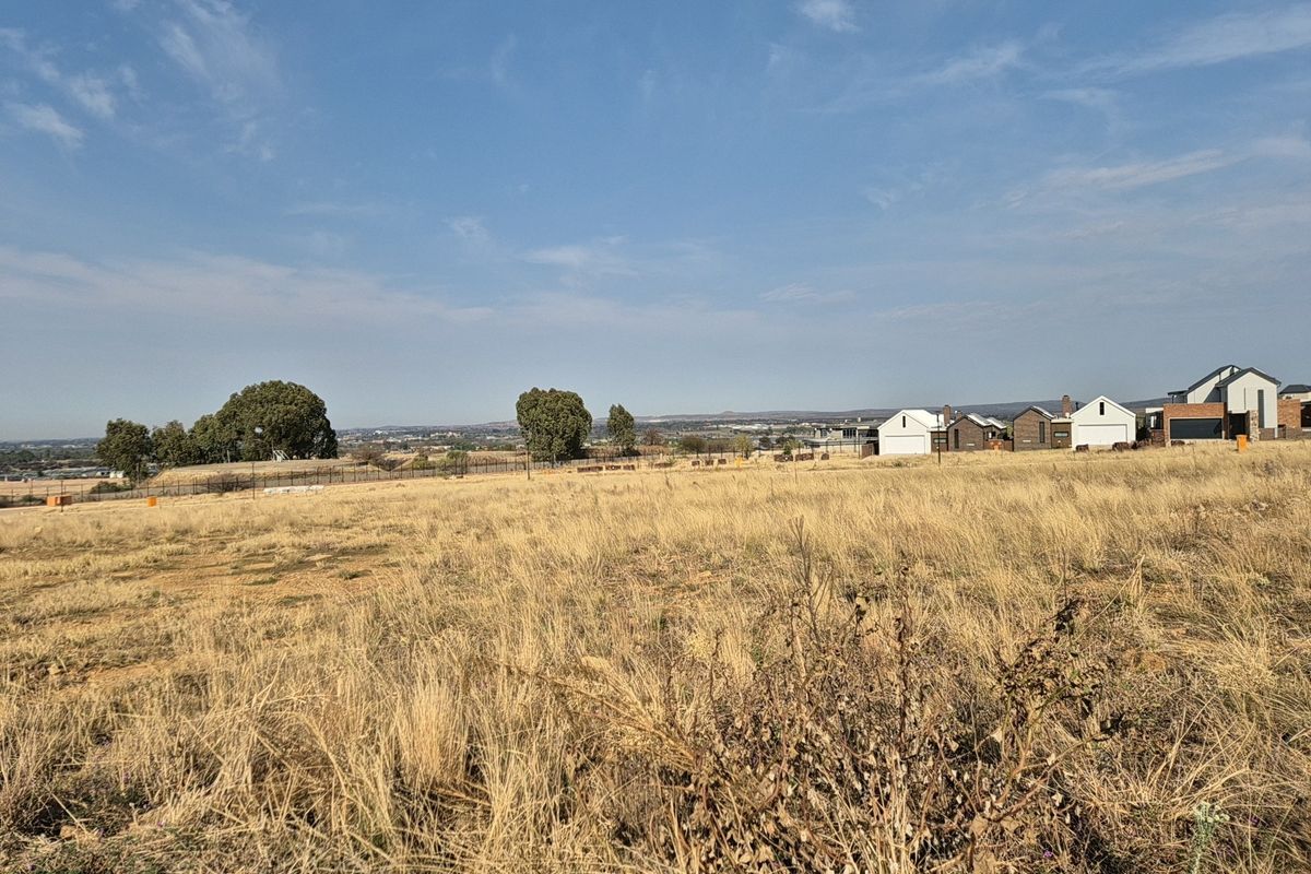A wide-angle view of a vacant residential plot featuring dry grass and surrounding neighborhood development in the distance.
