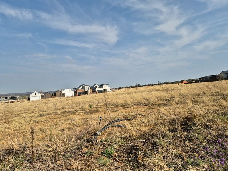 A view of a vacant residential plot featuring dry grass and visible utility conduits, with established modern homes in the background.