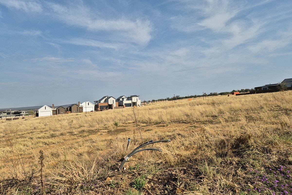 A view of a vacant residential plot featuring dry grass and visible utility conduits, with established modern homes in the background.