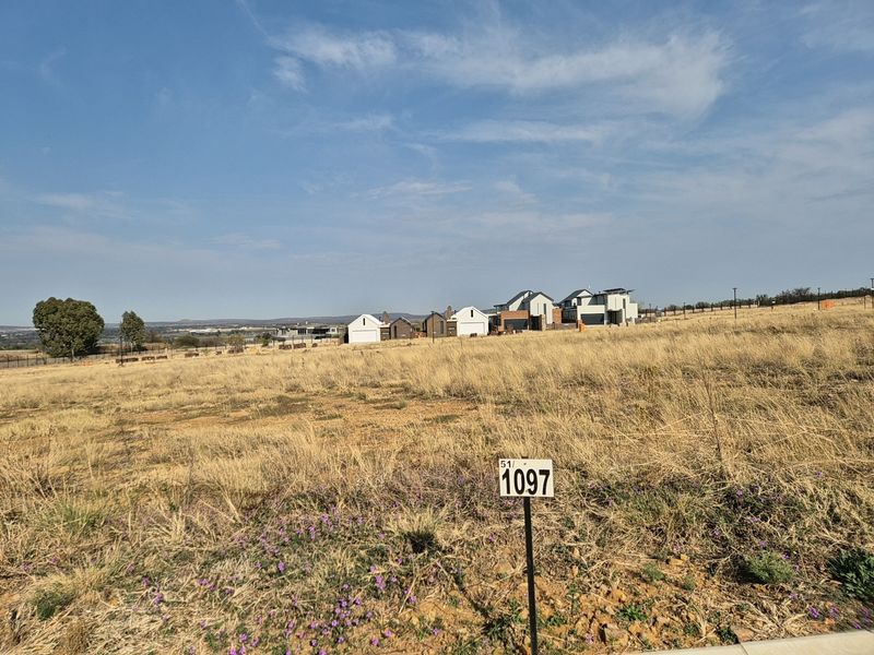 A view of a vacant residential plot featuring dry grass and a property boundary marker, with established homes visible in the background.