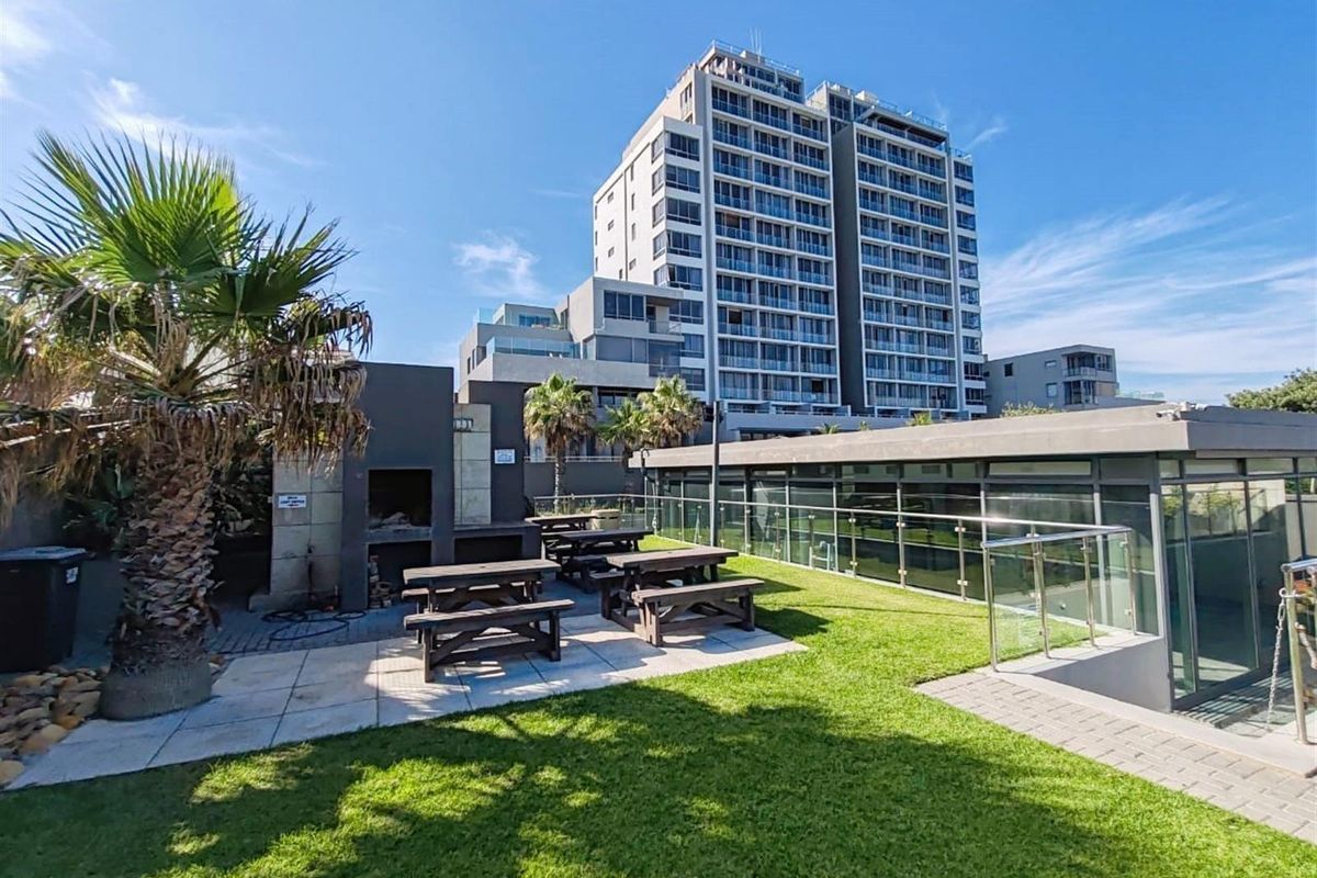 Outdoor communal entertainment area featuring a built-in braai, paved patio, and manicured lawn, set against the backdrop of a modern apartment building.