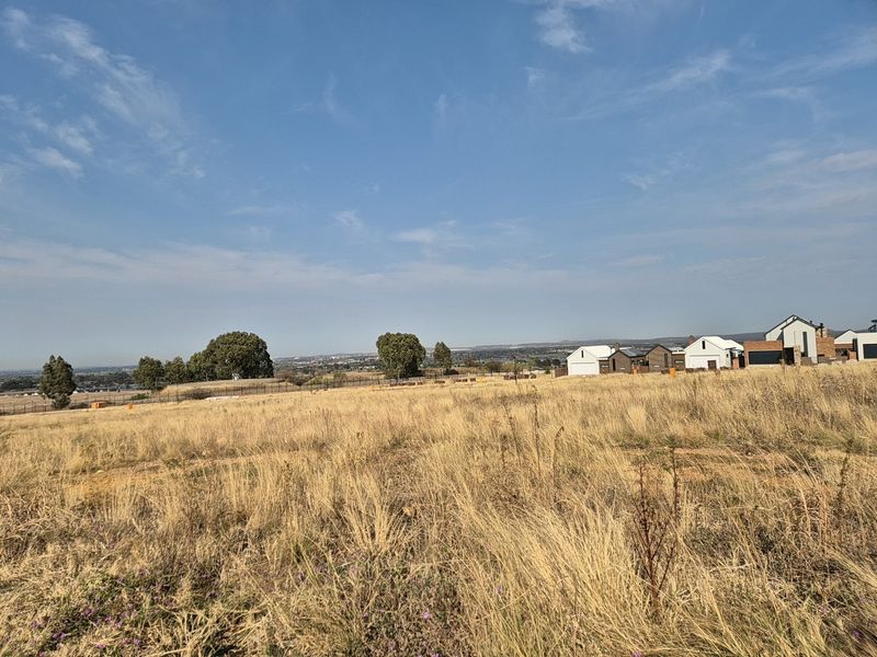 A wide-angle view of a vacant residential plot featuring dry grassland, perimeter fencing, and distant neighborhood houses under a clear blue sky.
