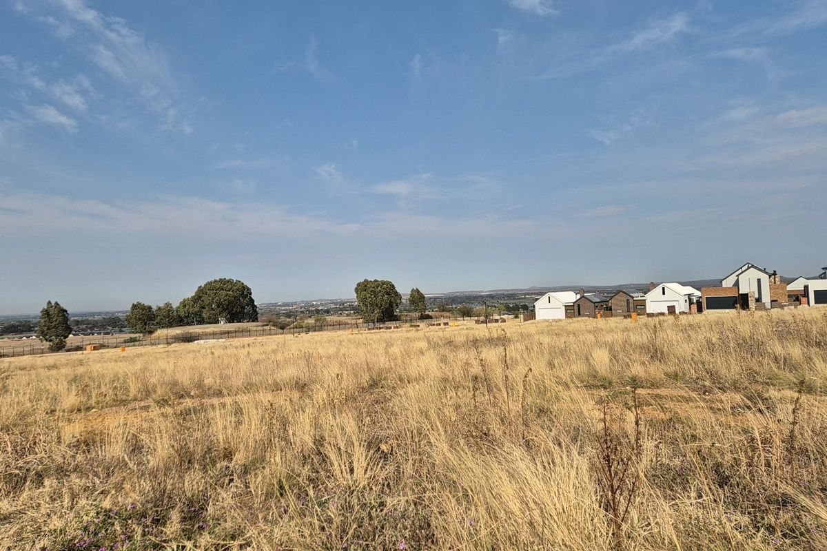 A wide-angle view of a vacant residential plot featuring dry grassland, perimeter fencing, and distant neighborhood houses under a clear blue sky.