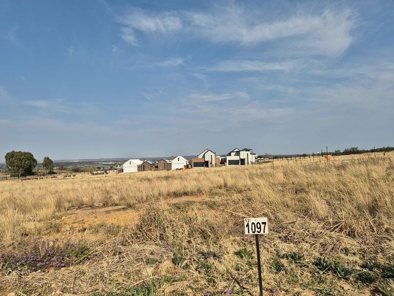 A view of a vacant residential plot featuring dry grass and surrounding neighborhood development in the distance.