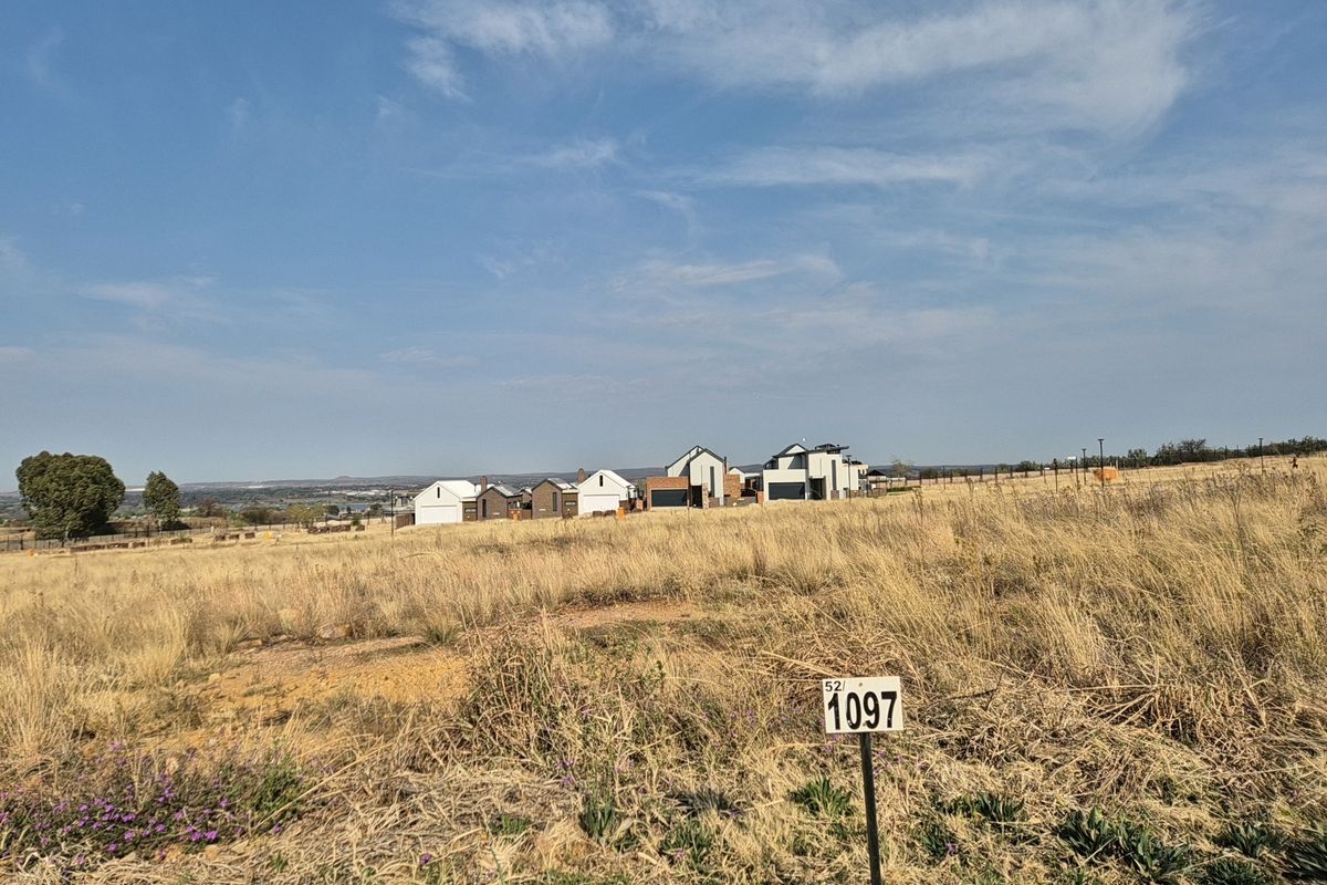 A view of a vacant residential plot featuring dry grass and surrounding neighborhood development in the distance.
