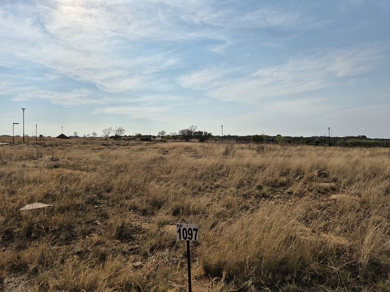 A vacant residential plot of land featuring natural grass coverage, boundary fencing, and existing street lighting infrastructure.