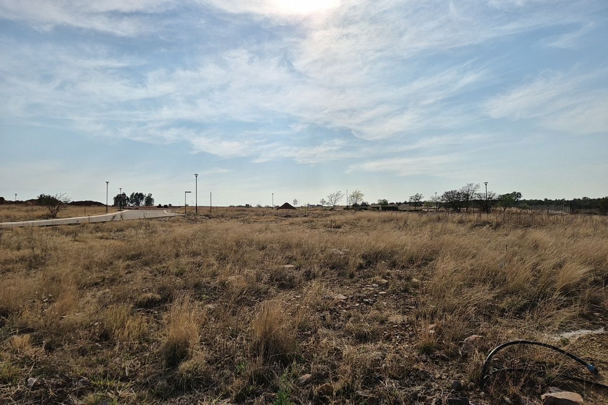 A vacant residential plot featuring natural grass cover, established street lighting, and direct access to a paved road in a developing estate.