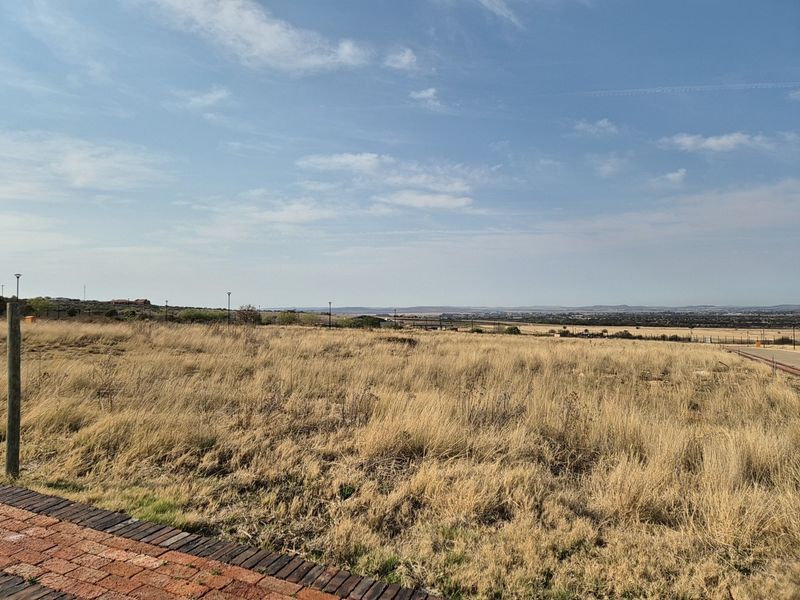 A vacant residential plot featuring dry grass, a paved verge, and distant landscape views, suitable for new construction.