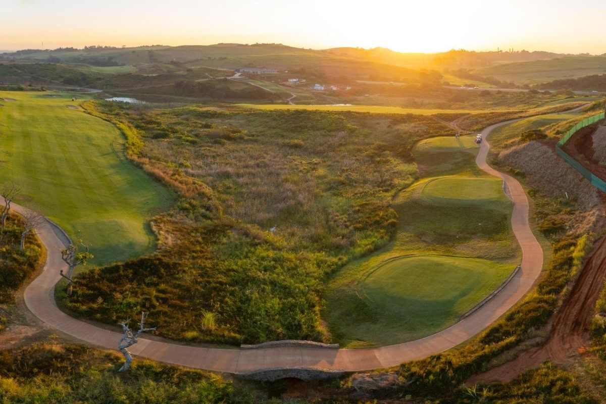 Aerial view of a golf course landscape featuring manicured fairways, paved cart paths, and a stone bridge at sunset.