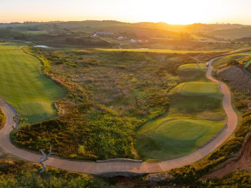 Aerial view of a golf course landscape featuring manicured fairways, paved cart paths, and a stone bridge at sunset.