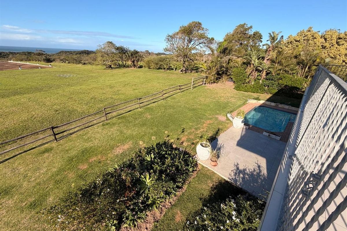Elevated view of the farm property featuring a swimming pool, paved patio area, expansive green lawn, and post-and-rail fencing with distant coastal views.