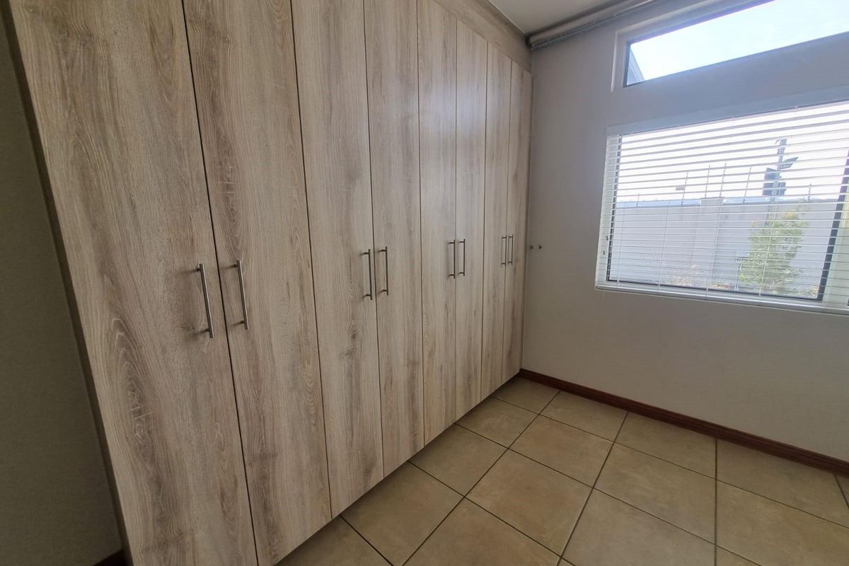 A clean, empty bedroom featuring extensive light wood-finish built-in cupboards, neutral tiled flooring, and a window with fitted blinds.