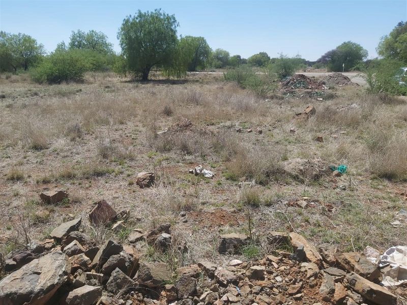 A view of a vacant residential plot featuring natural vegetation, rocky terrain, and scattered debris on the ground.