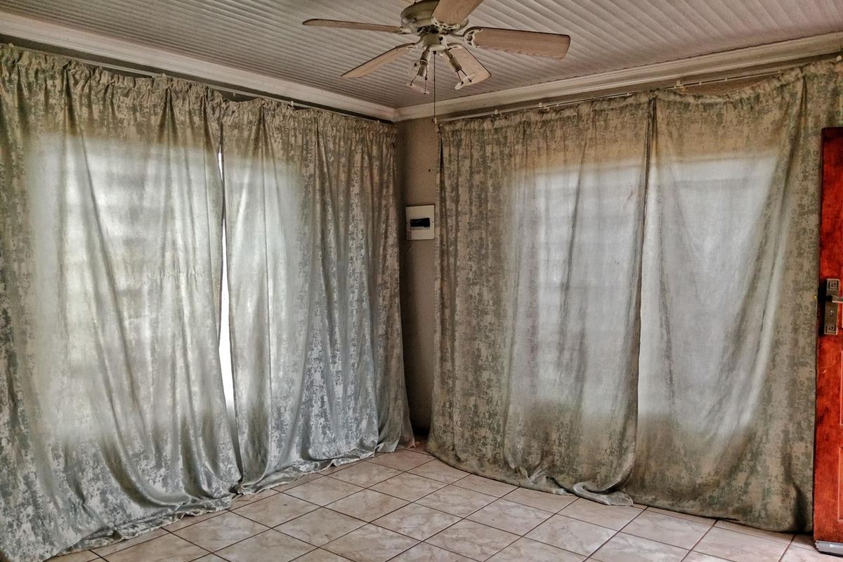 A living area featuring tiled flooring, a ceiling fan, and large windows covered by curtains, offering a blank canvas for renovation.