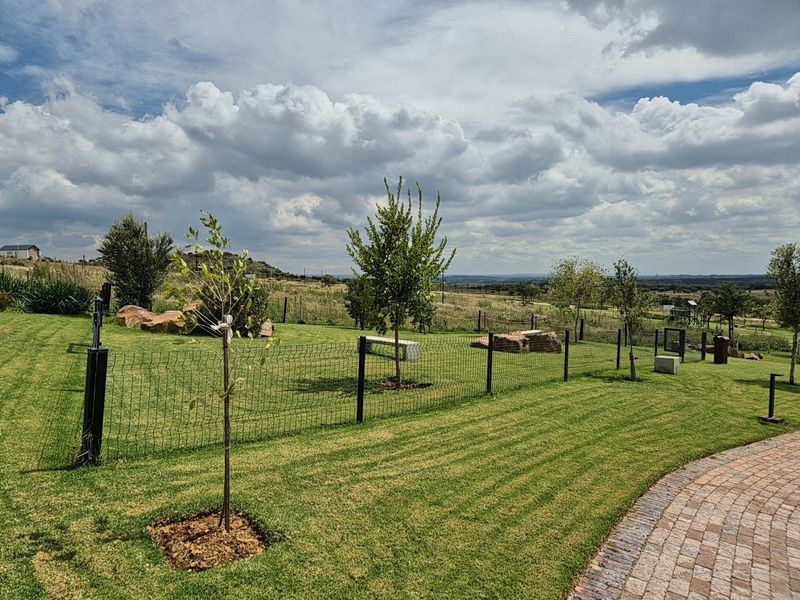 A well-maintained residential garden featuring a manicured lawn, paved walkway, and wire mesh fencing with open views of the surrounding landscape.