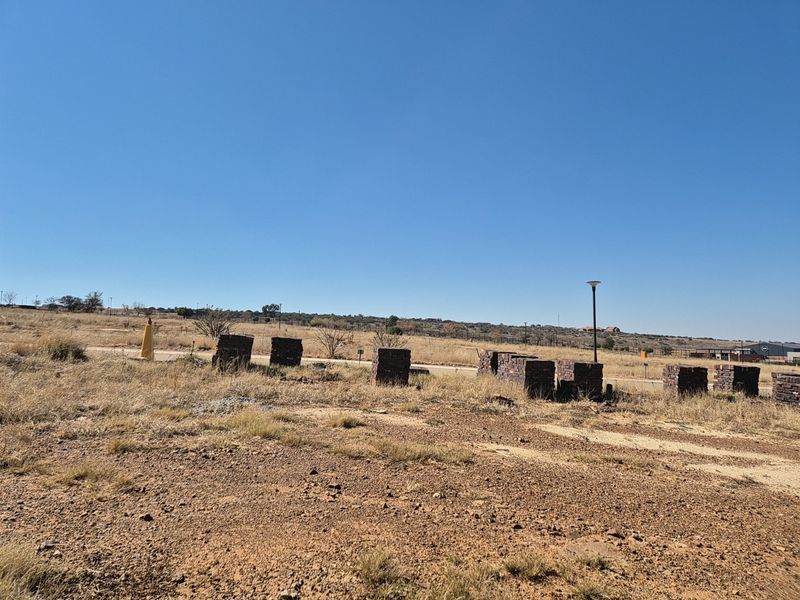 A vacant residential plot featuring dry grass, open terrain, and existing boundary markers under a clear blue sky.