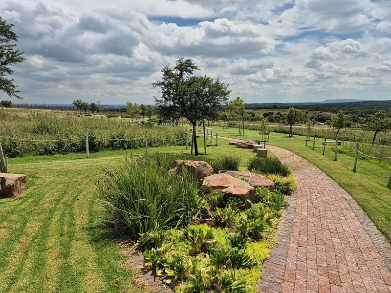 A well-maintained residential garden featuring a brick paved pathway, landscaped rock features, and open green space with established trees and rope fencing.