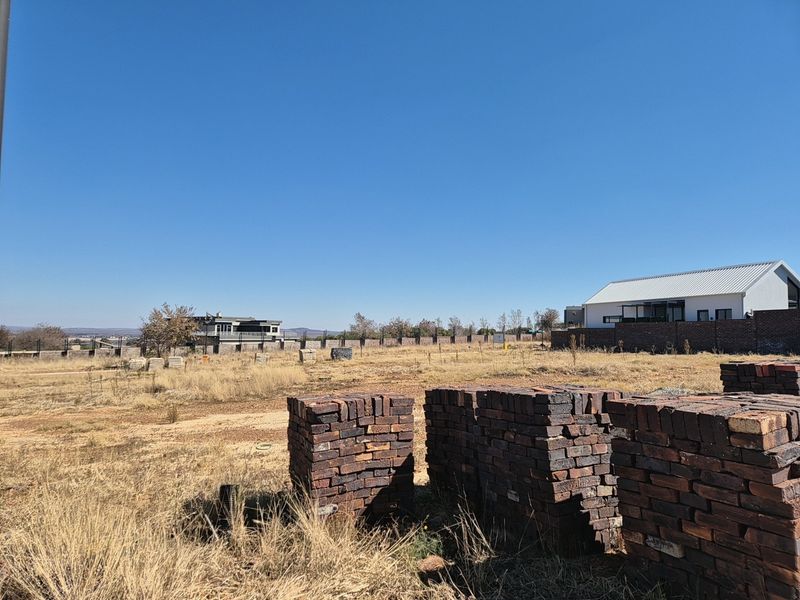 A vacant residential plot featuring stacks of construction bricks, dry grass, and a perimeter wall with neighboring properties visible in the background.