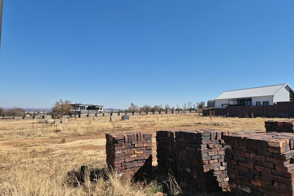 A vacant residential plot featuring stacks of construction bricks, dry grass, and a perimeter wall with neighboring properties visible in the background.