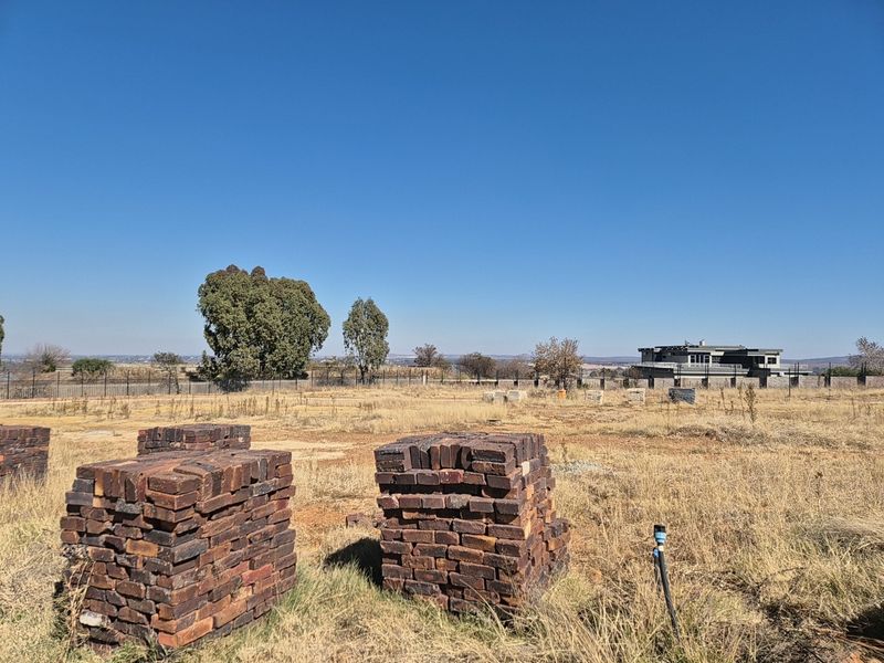A vacant residential plot featuring dry grass, perimeter fencing, and stacked building materials, offering an open landscape with distant neighborhood views.