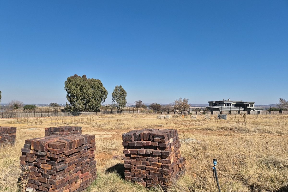 A vacant residential plot featuring dry grass, perimeter fencing, and stacked building materials, offering an open landscape with distant neighborhood views.