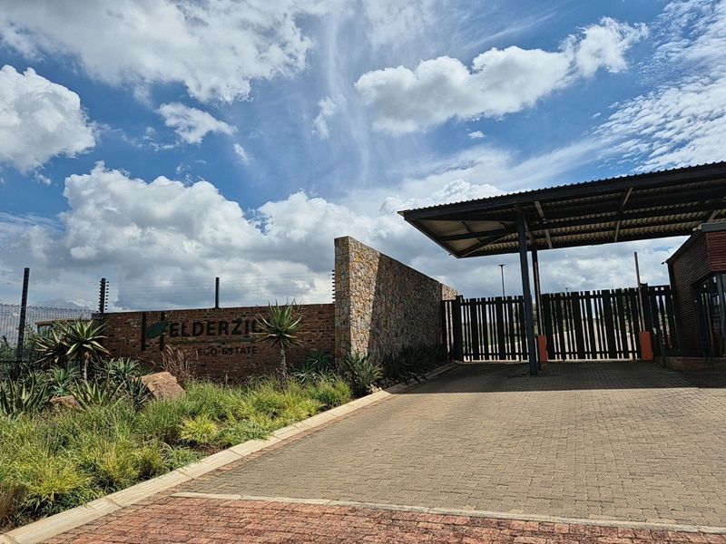 The secure entrance gate and stone-clad boundary wall of the Elderzio Eco Estate, featuring paved driveway access and integrated security fencing.