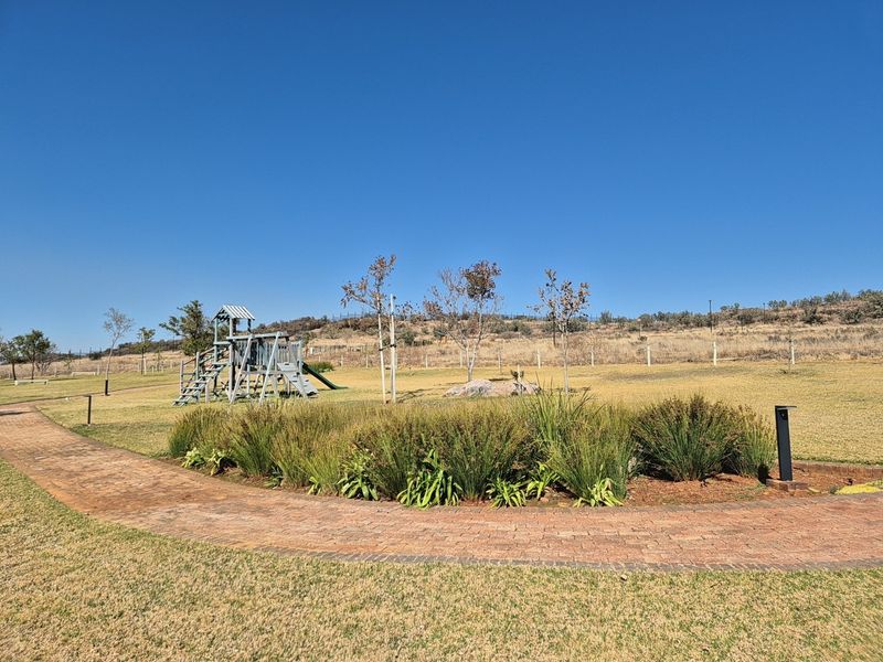 A well-maintained residential garden featuring a paved walkway, landscaped garden beds, and a playground set within an open, grassy area.