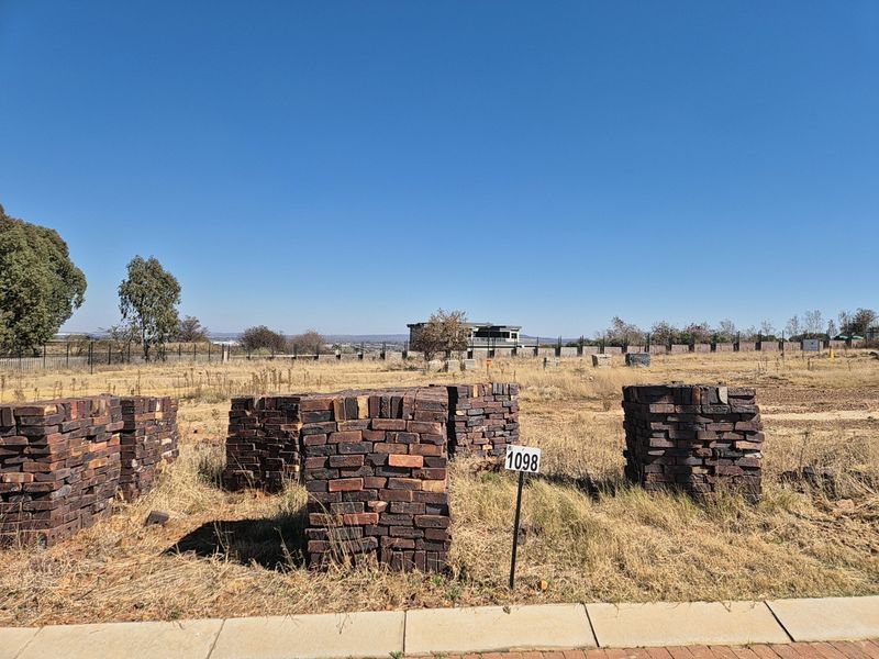 A vacant residential plot featuring dry grass and stacked brick piles, situated in a developed estate with perimeter fencing.