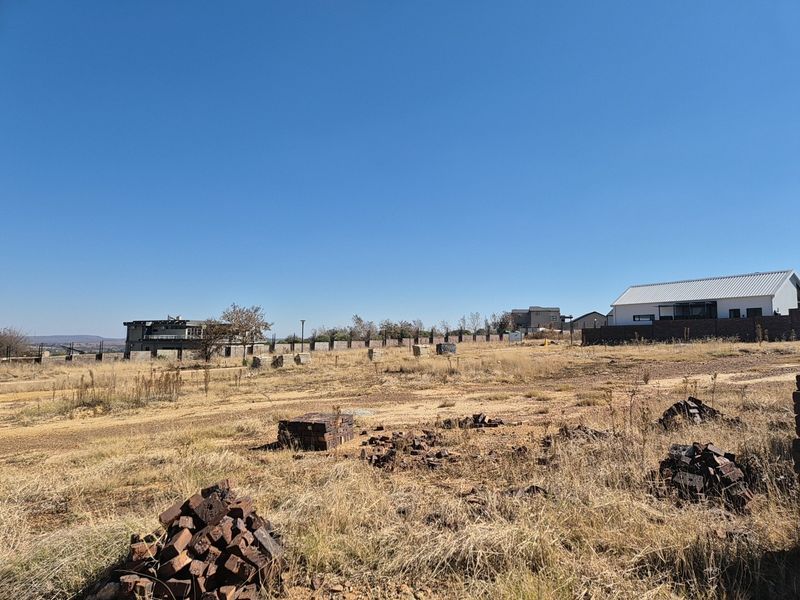 A view of a vacant residential plot featuring dry grass, scattered building materials, and surrounding neighborhood properties under a clear blue sky.