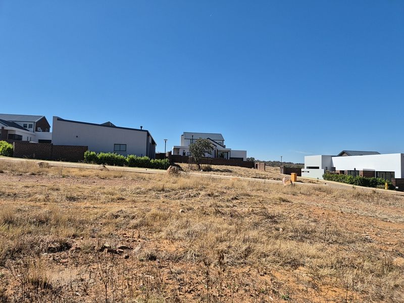 A vacant residential plot featuring natural dry grass terrain, situated in a developed estate with paved roads and modern neighboring homes.