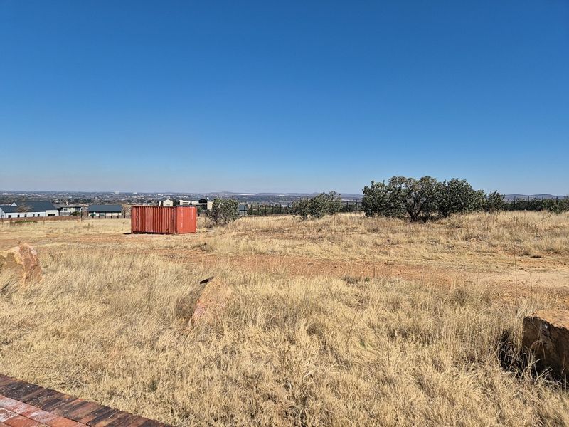 A wide-angle view of a vacant residential plot featuring dry grass, natural vegetation, and distant neighborhood views under a clear blue sky.