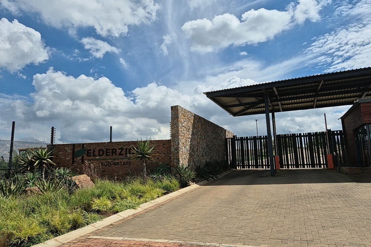 The entrance to Elderzio Eco Estate featuring a stone-clad boundary wall, electric fencing, and a secure gated access point with a paved driveway.
